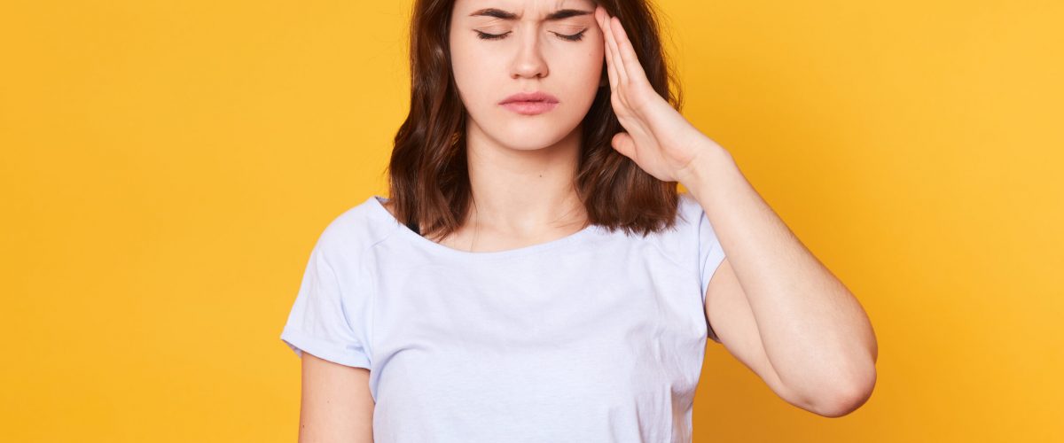 Close up portrasit of young brunette woman wearing white casual t shirt isolated over yellow background with hand on temple, has pain in head because stress, stands with closed eyes. Copy space.