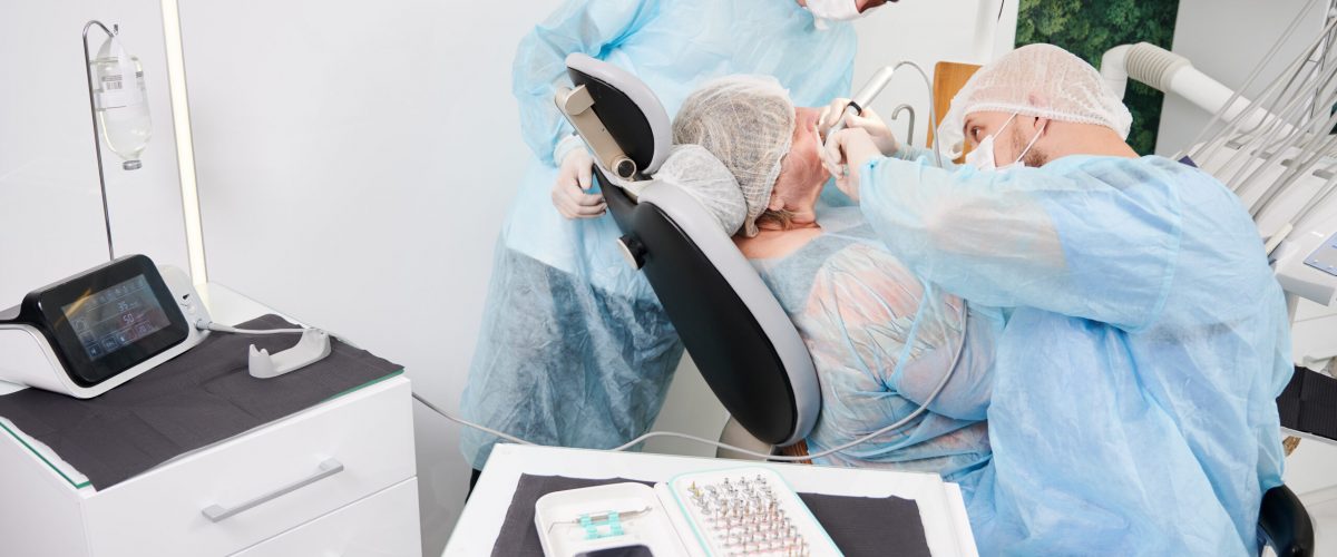 Implantology procedure with implant physiodispenser. Male dentist using dental implant machine while man colleague standing beside woman and observing the process. Concept of dental implant placement.