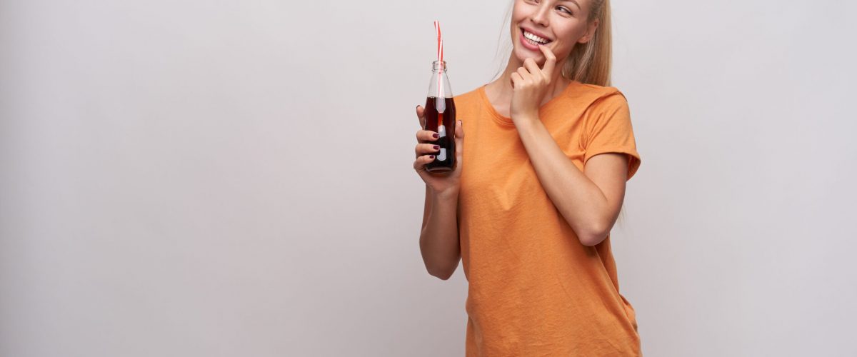 Indoor photo of cheerful young pretty blonde female in casual clothes holding bottle of soda in raised hand and keeping forefinger on underlip while looking aside with interest